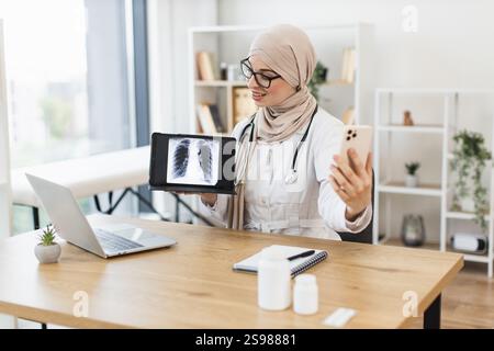 Médecin musulman féminin effectue un appel vidéo avec le patient, affichant une radiographie pulmonaire sur tablette. Stéthoscope professionnel d'âge moyen en cabinet médical. Banque D'Images
