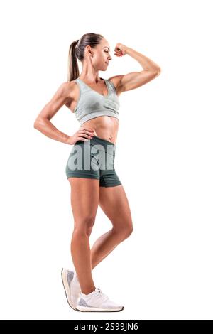 Femme athlétique dans les vêtements d'entraînement fléchissant les muscles du bras, posant avec confiance sur un fond blanc propre, mettant l'accent sur la force, la forme physique, la santé, un Banque D'Images
