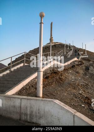 Un grand poteau blanc avec un globe sur le dessus se dresse sur une colline. La colline est couverte de rochers et il y a des escaliers qui y mènent. Le ciel est clair et bleu, Banque D'Images