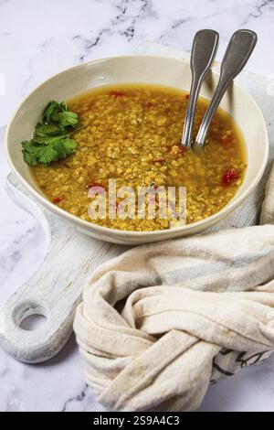 Un bol de soupe de lentilles rempli de saveurs chaudes et riches, garni d'herbes fraîches, accompagné de cuillères en argent, disposé sur un boa de coupe en bois léger Banque D'Images