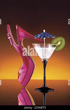 Une vue colorée de la chaussure rose à talons hauts d'une femme et d'un verre à cocktail, garni d'un parapluie en papier et d'une tranche de kiwi Banque D'Images