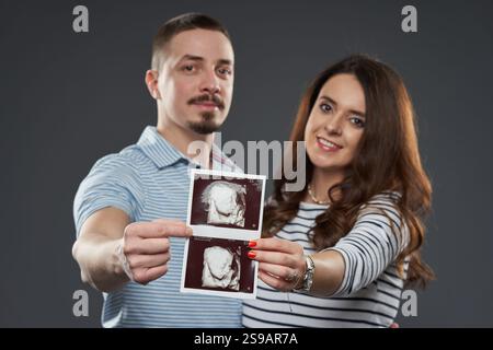 Attendre le couple tenant et montrant des images échographiques de leur bébé, souriant avec excitation et anticipation, isolé sur fond gris Banque D'Images