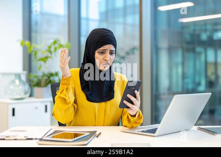 Femme lisant de mauvaises nouvelles en ligne au téléphone, employé de bureau en hijab tenant smartphone, regardant l'écran bouleversé et nerveux. Femme arabe au lieu de travail assis avec ordinateur portable à l'intérieur du bureau. Banque D'Images