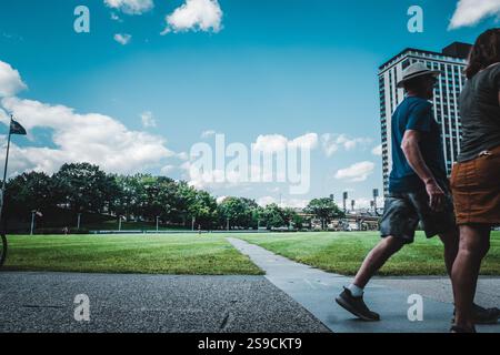 Un groupe d'individus se promène le long d'une passerelle pavée dans un parc sous un ciel bleu vif. L'herbe verte luxuriante et les bâtiments de la ville créent une atmosphère animée Banque D'Images
