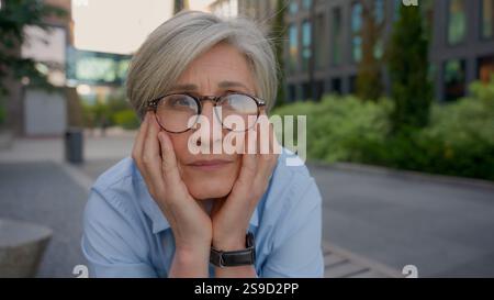 Caucasien vieille femme d'affaires senior âge moyen femme tristement réfléchie dame des années 50 dans les lunettes s'assoient à l'extérieur regardant la caméra triste expression mature Banque D'Images