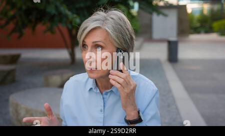 En colère folle femme d'affaires senior caucasienne vieille femme stressée femme argumente parler téléphone portable mécontent dame d'affaires mature sur la rue de la ville parler Banque D'Images