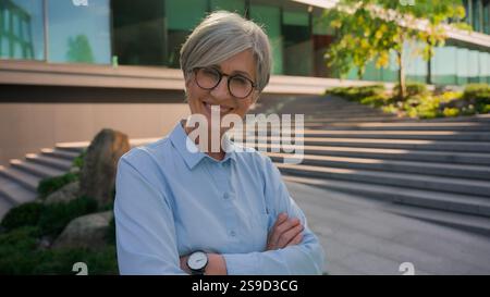 Portrait caucasien heureux confiant aux cheveux gris des années 60 femme d'affaires âgée femme d'âge moyen posant les mains croisées dans la ville à l'extérieur près du bureau urbain Banque D'Images