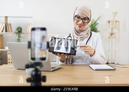 Jeune femme musulmane médecin enregistre la leçon en ligne avec le téléphone, expliquant le résultat de la radiographie des poumons. Elle utilise du matériel médical dans un bureau lumineux, en se concentrant sur l'éducation et la communication de soins de santé. Banque D'Images