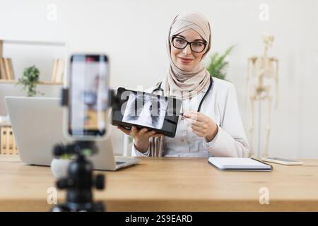 Jeune femme musulmane médecin enregistre la leçon en ligne avec le téléphone, expliquant le résultat de la radiographie des poumons. Elle utilise du matériel médical dans un bureau lumineux, en se concentrant sur l'éducation et la communication de soins de santé. Banque D'Images