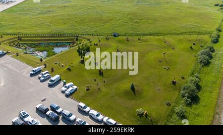 Vue aérienne d'un parc herbeux avec des sculptures et un petit étang, adjacent à un parking avec plusieurs voitures. Le paysage est entouré de f ouvert Banque D'Images