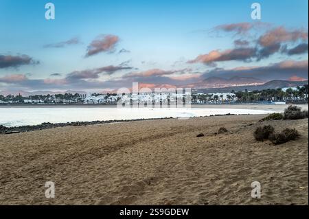Plage de sable à Los Pocillos, Lanzarote, avec des vagues calmes de l'océan, des montagnes lointaines, des palmiers et des bâtiments blancs sous un ciel coloré au coucher du soleil. Banque D'Images