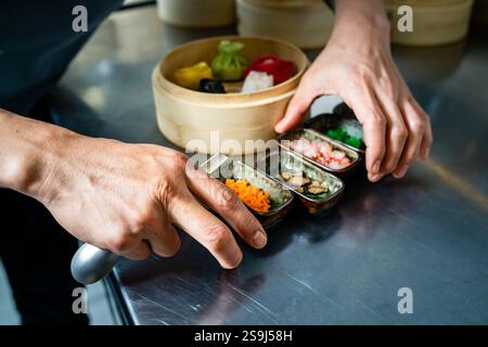 Gros plan d'un chef disposant de petits bols de condiments sur un comptoir en acier inoxydable, avec un cuiseur vapeur en bambou contenant des boulettes colorées dans le bac Banque D'Images