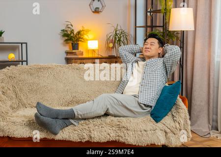 Heureux homme chinois d'âge moyen insouciant assis confortablement avec la main derrière la tête sur le canapé dans le salon à la maison. Souriant mec coréen se relaxant sur le canapé pensant regarder autour dans la chambre de l'appartement. Banque D'Images