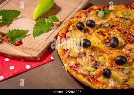 Pizza maison avec pepperoni, olives, mozzarella sur une planche de bois. Forme ronde, croûte épaisse, petites tranches de pepperoni, fines tranches d'olive, feuilles de menthe, Banque D'Images