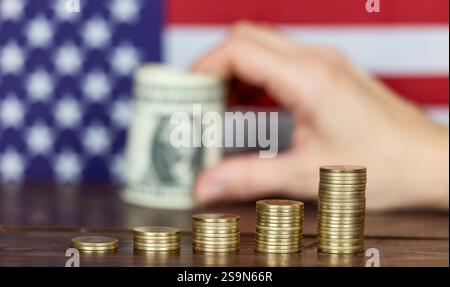 Une main tient des billets de banque en dollars derrière des piles de pièces de monnaie avec le drapeau des États-Unis en arrière-plan. Augmentation de l'inflation ou concept des taux d'intérêt. Banque D'Images