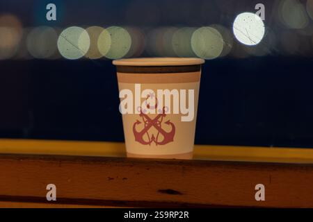 Un gobelet en papier avec un emblème maritime turc placé sur une surface en bois la nuit. Les lumières floues de la ville créent un effet bokeh en arrière-plan. IBB, 2025 Banque D'Images