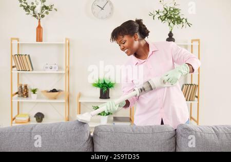 Femme souriante heureuse nettoyant canapé avec aspirateur dans le salon à la maison. Banque D'Images
