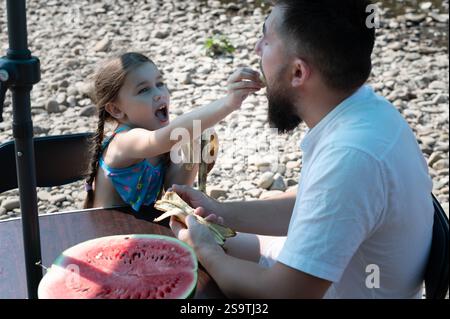 Une scène réconfortante d'une fille et son père partageant un pique-nique au bord d'une rivière rocheuse. Elle lui nourrit ludique d'une banane tandis qu'une pastèque se trouve à proximité. Banque D'Images