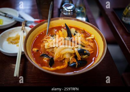 Jjamppong Udon dans un bol de soupe rouge avec une cuillère dedans. Le bol est sur une table. Il y a des baguettes sur la table Banque D'Images