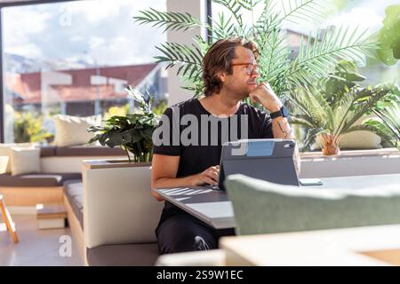 Un homme d'affaires hipster est assis à une table avec une tablette et a une expression réfléchie. Banque D'Images