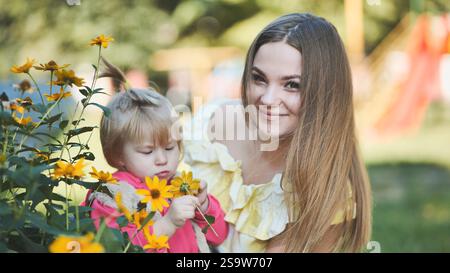 Mère souriante et enfant se liant tout en ramassant des fleurs sauvages jaune vif dans un cadre luxuriant de parc d'été Banque D'Images