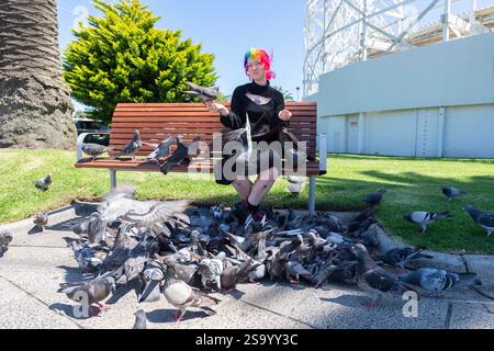 Une fille aux cheveux multicolores est assise sur un banc de parc, tient un pigeon dans sa main et nourrit des pigeons dans O'Donnell Gardens, un parc à St Kilda, Melbourne Banque D'Images