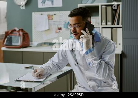 Professionnel de la santé écrivant des notes tout en parlant au téléphone dans un cadre de bureau ayant des lunettes et portant un manteau blanc tout en travaillant au bureau Banque D'Images