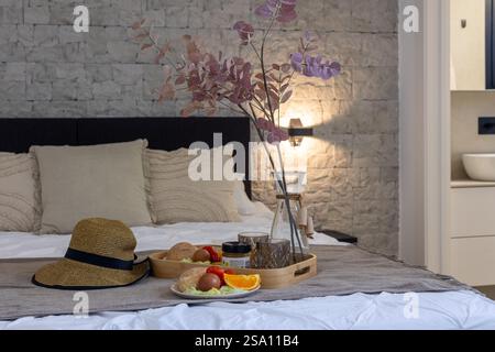 Une configuration de lit confortable avec un plateau de petit déjeuner avec de la nourriture, un chapeau de paille, un vase décoratif, et un mur de pierre rustique en toile de fond. Banque D'Images