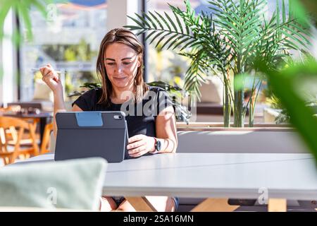 Une femme est assise à une table travaillant avec une tablette, elle a une expression heureuse et détendue. Banque D'Images