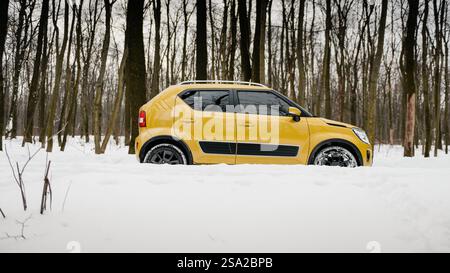 Vue latérale de Suzuki Ignis dans la neige profonde. Véhicule multisegment couleur moutarde conduite dans une forêt hivernale enneigée. Banque D'Images