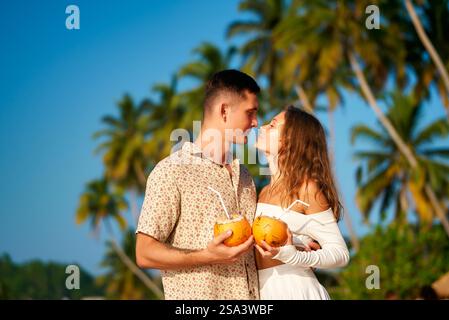 Couple romantique sur la plage tropicale, tenant des noix de coco, partageant un moment affectueux. Chemise à motifs homme, robe blanche femme, profitant du coucher du soleil. Amour Banque D'Images