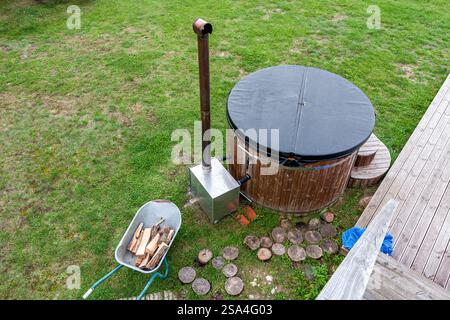 Un bain à remous extérieur en bois avec un chauffe-bois, à côté d'une pelouse et d'une terrasse. Une brouette de bois de chauffage se trouve à proximité, et le lieu de détente est situé à l'intérieur Banque D'Images
