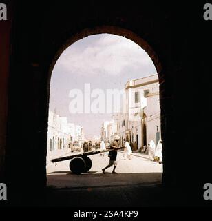 Blick durch einen Torbogen auf die Straße einer Medina, ein Lastenfahrer im Vordergrund, Marokko 1959. Vue à travers une arche sur la rue d'une médina, un chauffeur de marchandises au premier plan, Maroc 1959. Banque D'Images