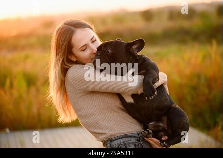 Femme tient un bouledogue français dans ses bras, embrasse le chien, le regarde avec amour au coucher du soleil. Le concept d'amitié, confiance, amour, sans enfant. Banque D'Images
