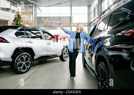 Femme prenant un selfie avec sa nouvelle voiture dans la concession pendant les vacances, agitant à la caméra Banque D'Images