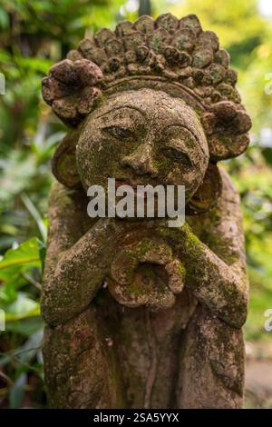 Une statue sereine en pierre balinaise couverte de mousse entourée d'une végétation luxuriante, reflétant l'essence tranquille et artistique des artisans traditionnels de Bali Banque D'Images