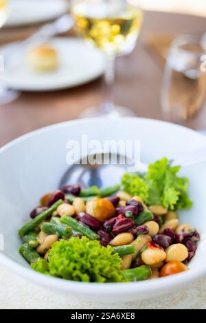 Une salade de haricots frais mélangée servie dans un bol blanc, garnie de laitue verte et de tomates cerises. Un verre de vin blanc et un cadre de table complètent le restaurant Banque D'Images