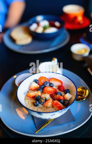 Flocons d'avoine chauds servis dans un bol blanc et bleu, garnis de fraises fraîches, de myrtilles et de crème sur assiette bleue. Banque D'Images