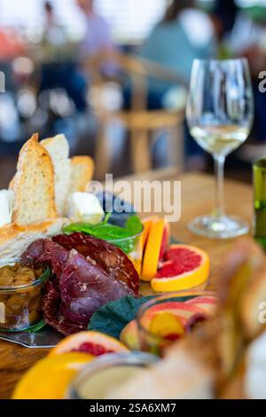 Une sélection vibrante de charcuteries, de fromage, de tranches de fruits frais et de pain, accompagnée d'un verre de vin blanc, le tout dans un cadre légèrement flou et détendu Banque D'Images