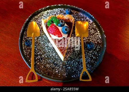 Vue en angle élevé une tranche de délicieux cheesecake maison garni de crème blanche, de fraise et de myrtille dans une plaque de céramique noire sur une table en bois Banque D'Images