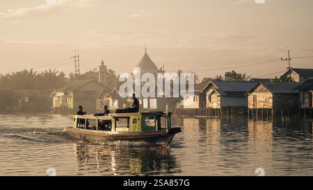 Un bateau Kelotok transportant des touristes le long de la rivière Martapura avec un village traditionnel et une mosquée en arrière-plan Banque D'Images