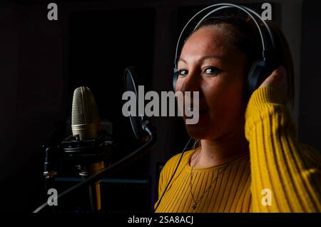 Chanteuse enregistrant une piste musicale dans un studio professionnel sombre à l'aide d'un microphone et d'un casque de haute qualité Banque D'Images
