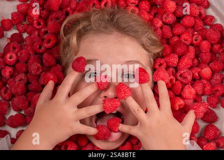 Visage drôle d'enfants près du fond de framboise. Enfant mignon mange des framboises des doigts. Framboise crue fraîche près du visage des enfants. Macro Raspberri. Vue de dessus de Banque D'Images