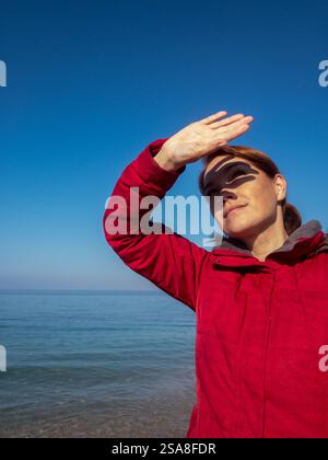 Femme en manteau rouge sur la plage de la mer regardant près de son visage à la main se cachant de la lumière du soleil Banque D'Images