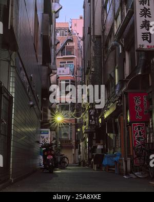 Vue tôt le matin d'une ruelle vide entre de grands bâtiments dans le centre de Tokyo, avec plusieurs panneaux, vélos, boutiques et une moto visible Banque D'Images