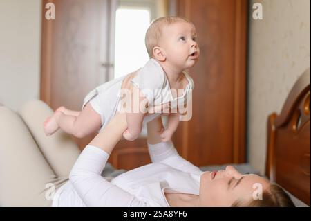 Mère aimante se trouve sur le lit et soulève son bébé mignon dans ses bras au-dessus d'elle. Femme regarde l'enfant avec amour et montre la protection. Banque D'Images