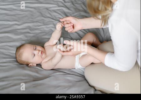Maman est assise sur le lit et joue avec le nourrisson allongé sur le lit, tenant tendrement ses mains. La femme aime la maternité. Concept de famille, amour, prendre soin d'un enfant, Banque D'Images