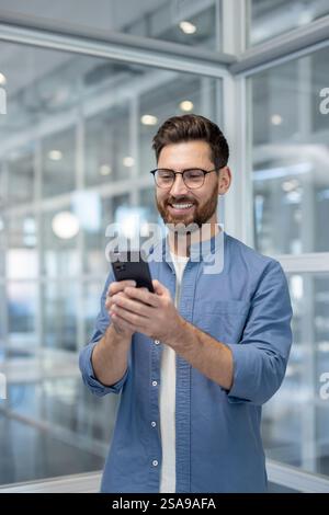 Plan vertical, homme heureux réussi debout à l'intérieur du bureau à l'aide du téléphone. Homme d'affaires tapant un message texte et discutant sur les médias sociaux. Banque D'Images