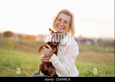 Femme sourit et tient amoureusement petit chien russe Toy Terrier aux cheveux lisses dans ses bras à l'extérieur. Les gens et les chiens. Journée mondiale du chien. Banque D'Images