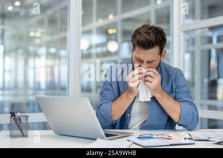 Homme malade souffrant au travail, éternuant. L'ouvrier a le nez qui coule et souffle froid de la morve dans une serviette. Travailler avec un ordinateur portable au bureau. Banque D'Images